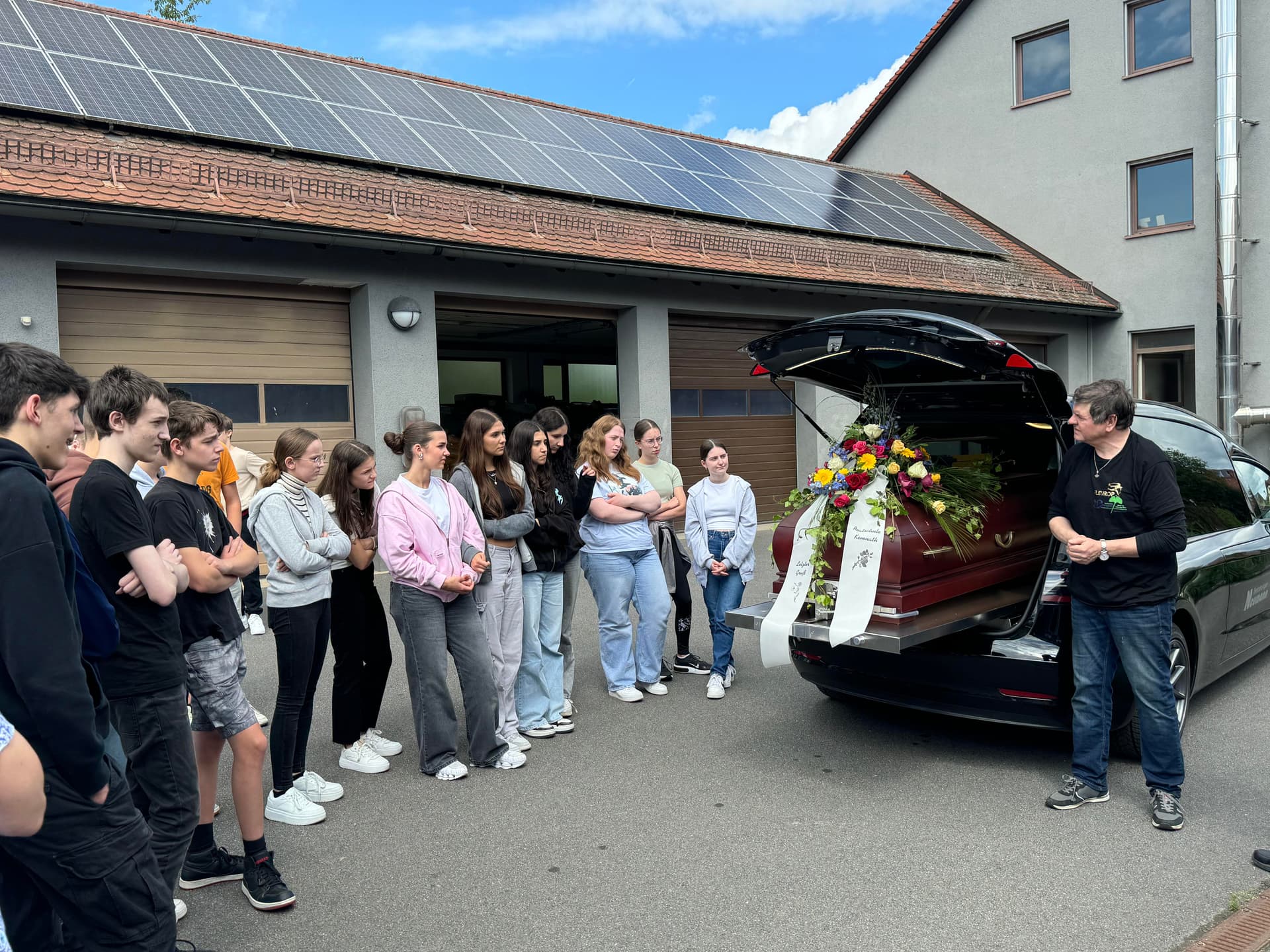 Schulklasse steht vor Leichenwagen mit Blumenschmuck